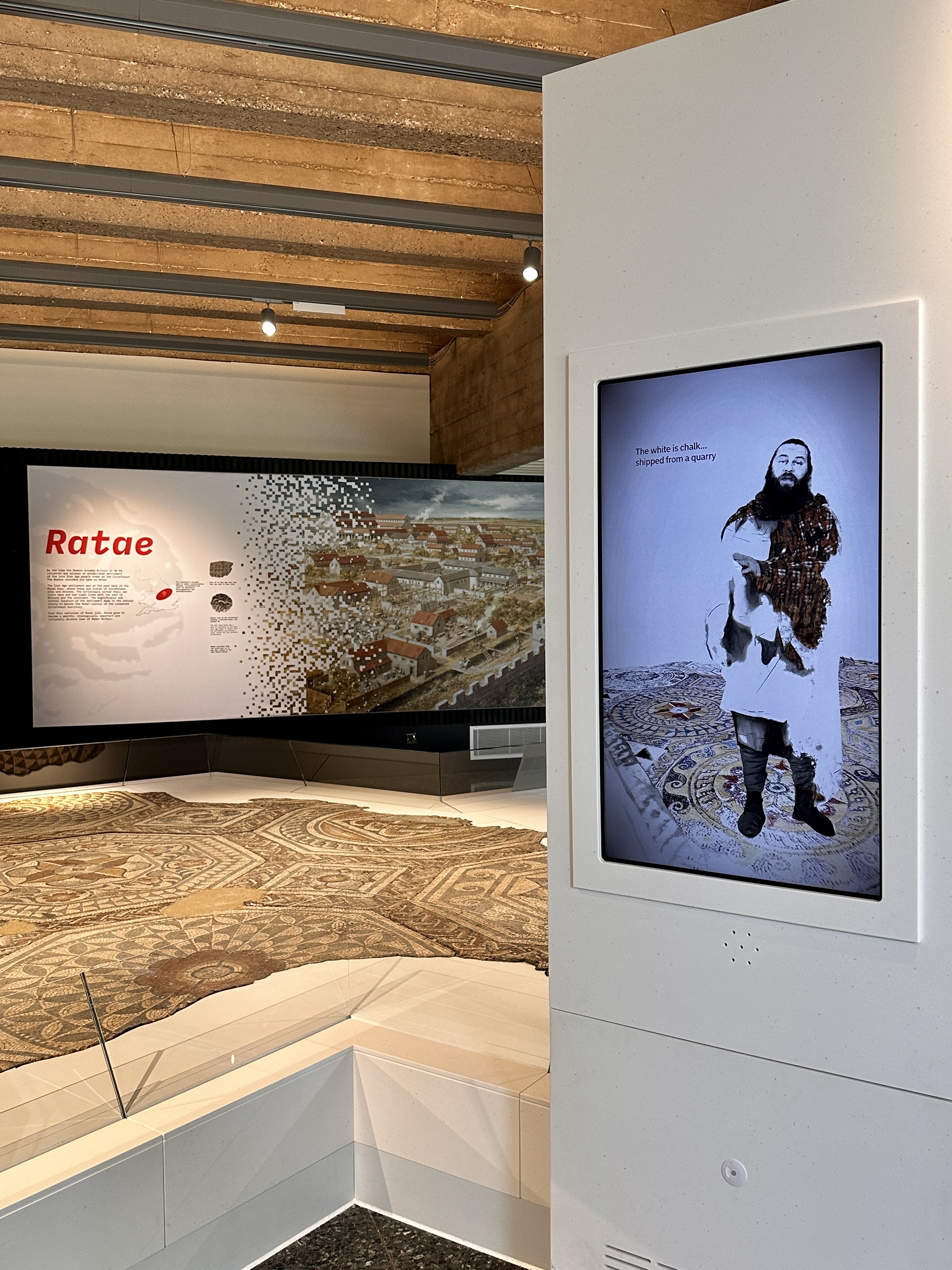 A gallery space with a roman mosaic on the ground and a video playing nearby of an ancient roman mosaic maker