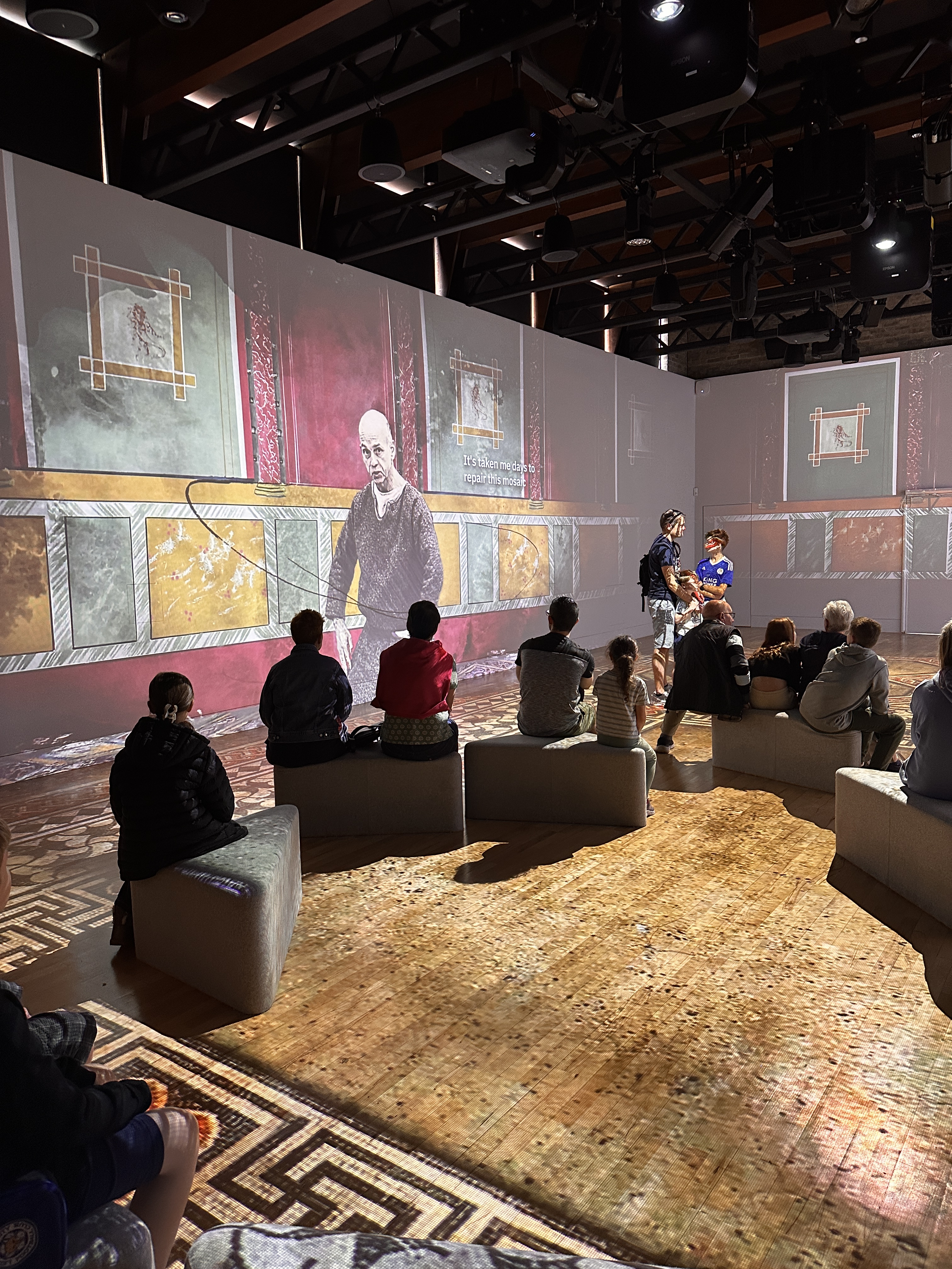 People sit in a large open room surrounded by wall sized projections displaying ancient roman life